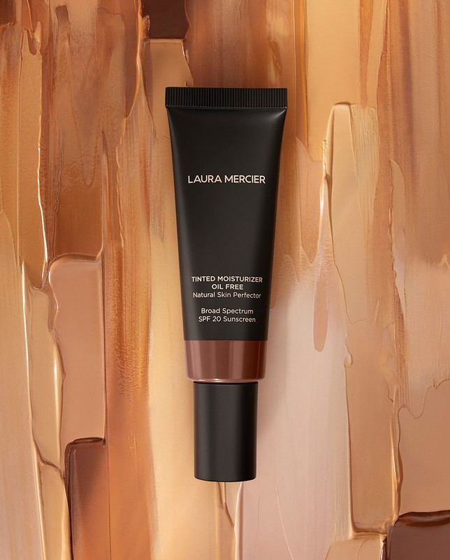 Laura Mercier tinted moisturizer tube on a background of textured beige, tan, and brown foundation makeup smears.