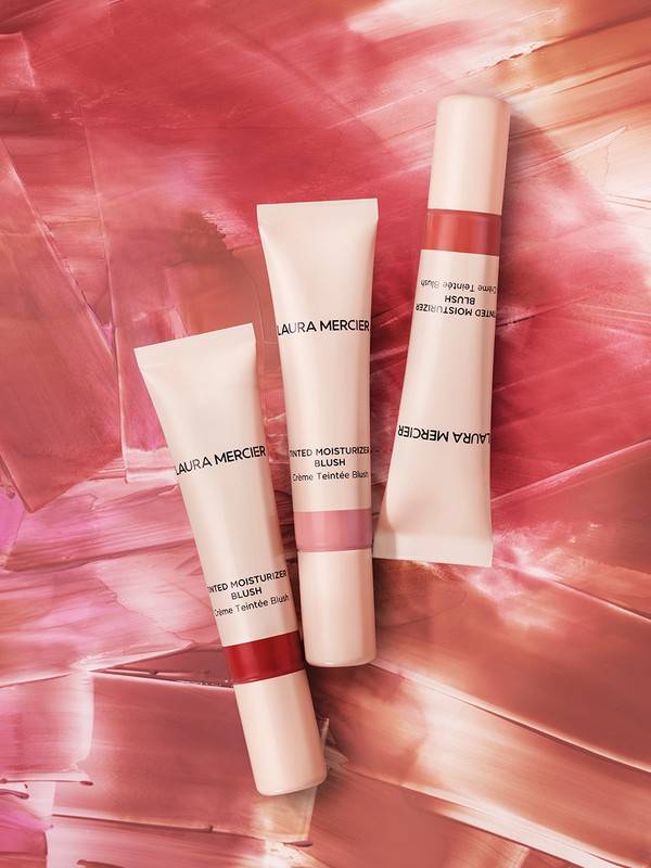 Three Laura Mercier blush tubes arranged against a textured, abstract background in shades of pink, peach, and white hues.