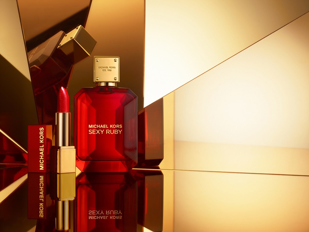 Still life shot of Michael Kors 'Sexy Ruby' perfume bottle with a lipstick tube; set against a background of geometric gold surfaces.