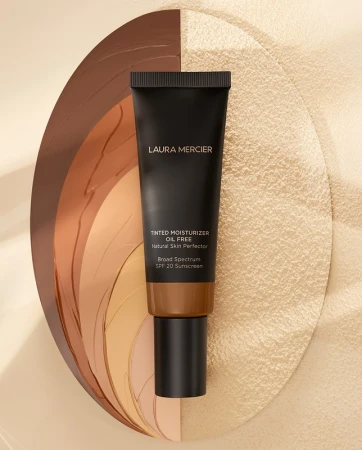 A black tube of Laura Mercier tinted moisturizer resting on a textured oval backdrop displaying swatches of different foundation shades.