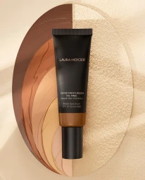 A black tube of Laura Mercier tinted moisturizer resting on a textured oval backdrop displaying swatches of different foundation shades.