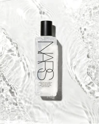 Overhead product shot of a clear rectangular NARS makeup removing water bottle resting in rippling water with bright lighting.