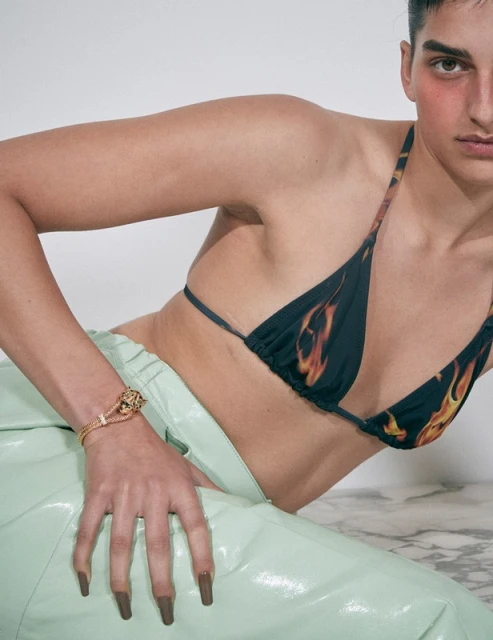 Female model poses in a photo studio wearing a flame-print bikini top, shiny green pants, and a gold bracelet on a white marble surface.