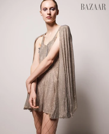 Model wearing a shimmering metallic chainmail dress with a matching cape against a neutral studio background