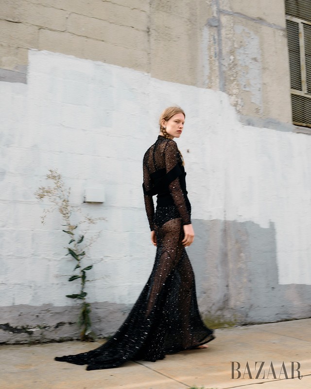 Fashion editorial shot: A young woman in a long black sparkling dress walks near a white wall, with Bazaar logo visible in the frame.