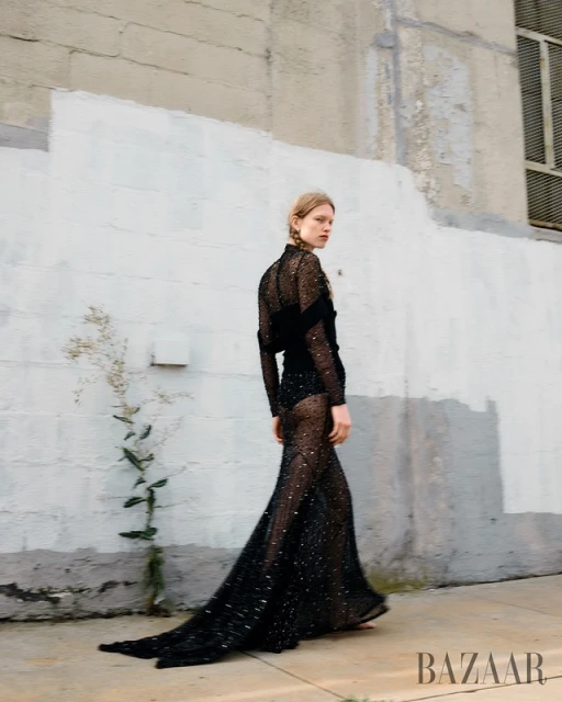 Fashion editorial shot: A young woman in a long black sparkling dress walks near a white wall, with Bazaar logo visible in the frame.