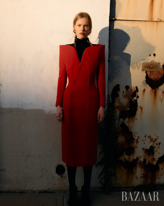 Editorial fashion shot of a woman in a red dress with padded shoulders standing in front of a textured wall. Fashion and style.