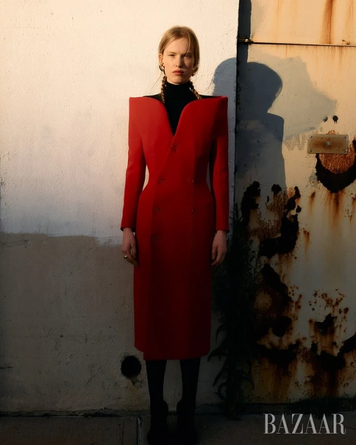 Editorial fashion shot of a woman in a red dress with padded shoulders standing in front of a textured wall. Fashion and style.