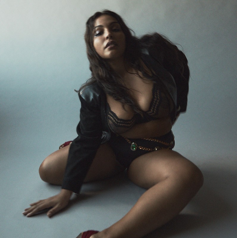 Portrait of a woman with long brunette hair, wearing black lingerie, a jacket, and gold chain belt, posing against a grey background.
