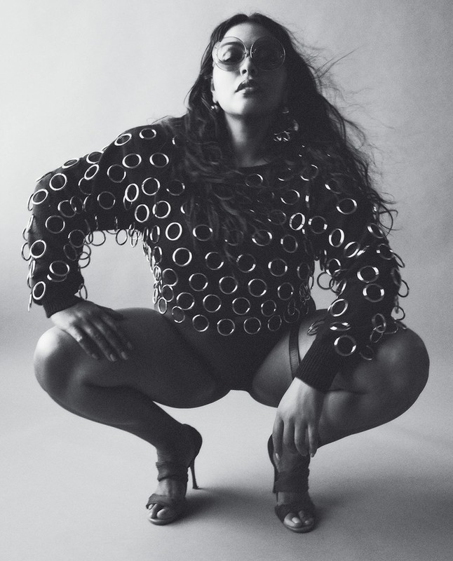 Monochrome portrait of a woman squatting in circular-patterned top, with round sunglasses and strappy heeled sandals in a photo studio.