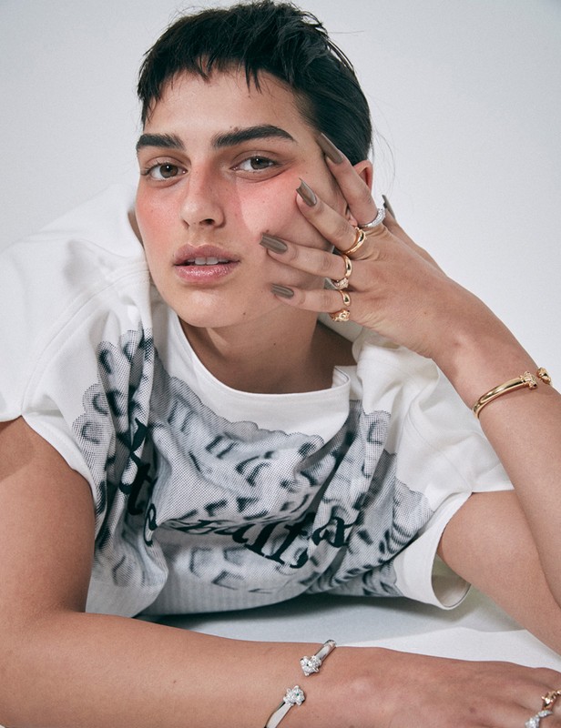 A person with short dark hair and brown eyes is lying down with their hands near their face and wearing bracelets and rings.