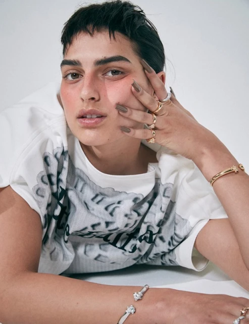 A person with short dark hair and brown eyes is lying down with their hands near their face and wearing bracelets and rings.