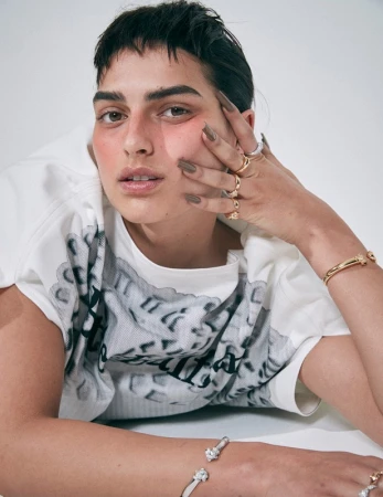 A person with short dark hair and brown eyes is lying down with their hands near their face and wearing bracelets and rings.