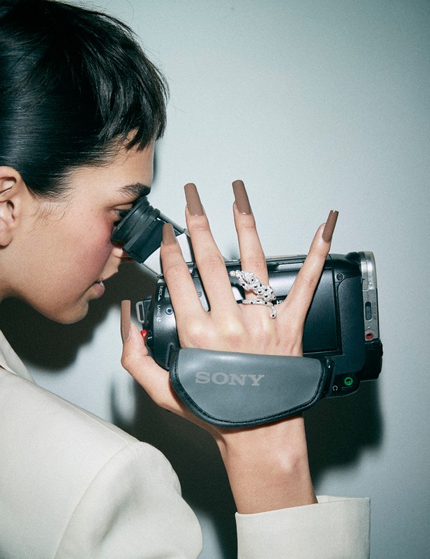 Woman films with a camcorder, displaying long brown nails, an animal ring, and a white jacket, set against a neutral backdrop.