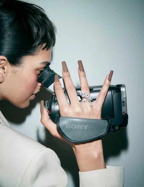 Woman films with a camcorder, displaying long brown nails, an animal ring, and a white jacket, set against a neutral backdrop.