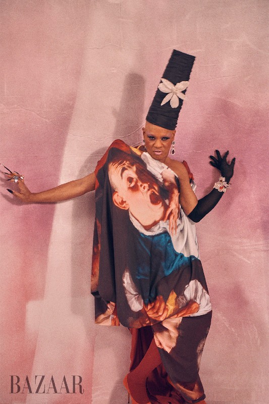 Unique fashion portrait of a person in dramatic makeup, a tall hat, and a gown with a face print, posing against a pink backdrop.