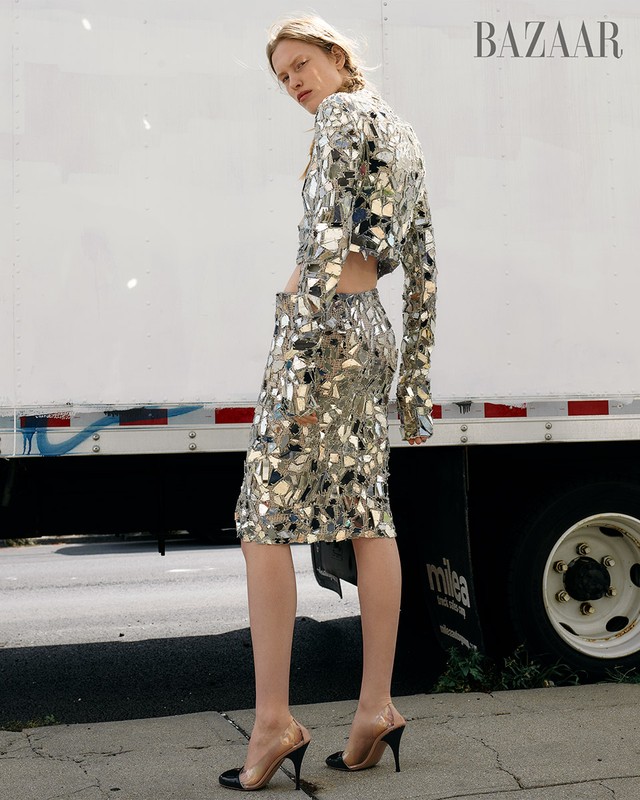 Fashion editorial shot of a fair-skinned model in a mirrored skirt and top, outdoors, with a white truck in the background.