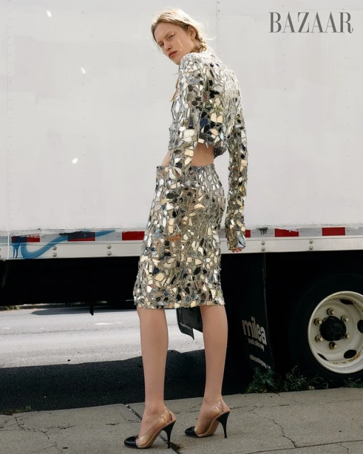 Fashion editorial shot of a fair-skinned model in a mirrored skirt and top, outdoors, with a white truck in the background.