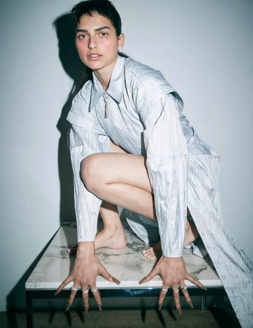 Attractive brunette woman in shiny futuristic outfit crouches on a marble table, gazing intently at the viewer in a fashion editorial.