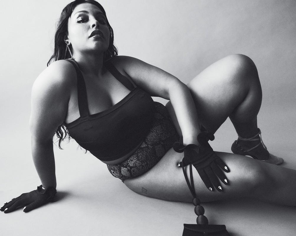 A woman in lingerie reclines in a studio, wearing gloves and looking up at the camera against a gradient neutral background.