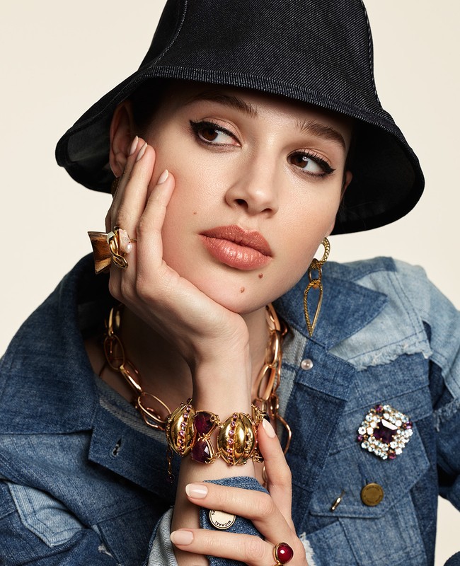 Close-up fashion shot of a young adult female model wearing a denim jacket, gold jewelry, and a dark hat, posing against a neutral background.