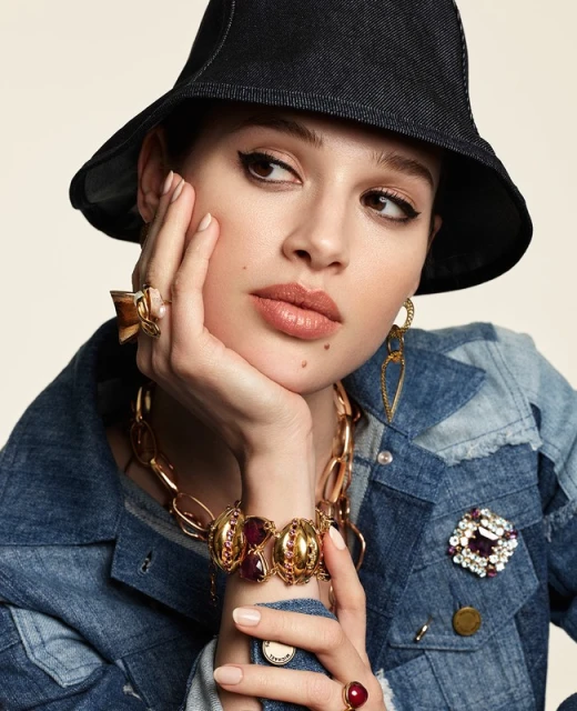 Close-up fashion shot of a young adult female model wearing a denim jacket, gold jewelry, and a dark hat, posing against a neutral background.