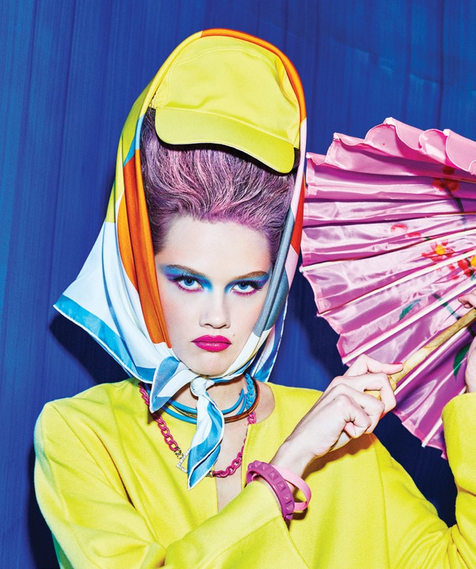 Woman with colorful makeup and headscarf holds pink parasol against a blue backdrop in a beauty and fashion editorial style shot.