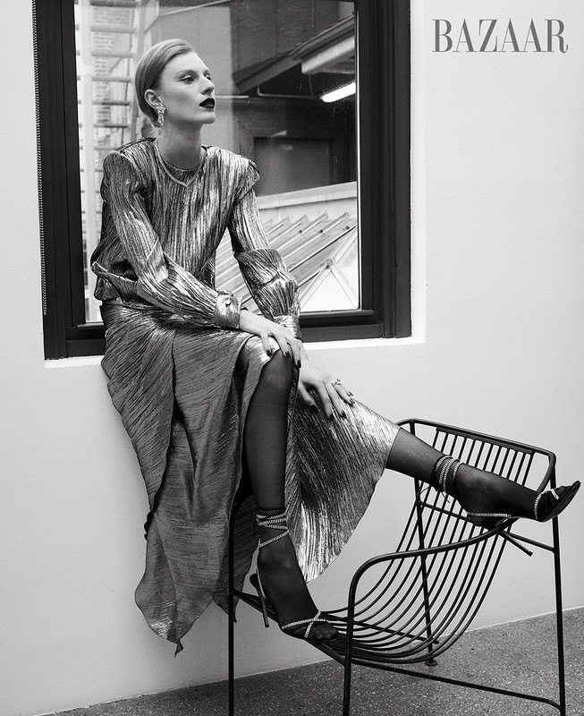 A woman poses in a metallic outfit in a black and white fashion shot, seated by a window in a long-sleeved top and matching skirt.
