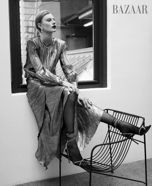 A woman poses in a metallic outfit in a black and white fashion shot, seated by a window in a long-sleeved top and matching skirt.
