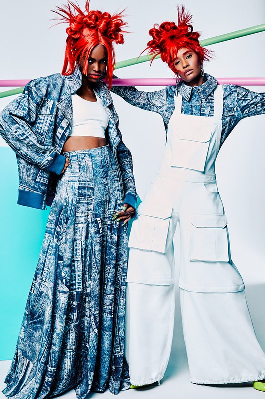Two Black female fashion models pose in a photo studio wearing avant-garde clothing in white and blue, colorful hair, and bold makeup.