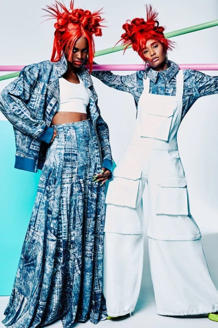 Two Black female fashion models pose in a photo studio wearing avant-garde clothing in white and blue, colorful hair, and bold makeup.