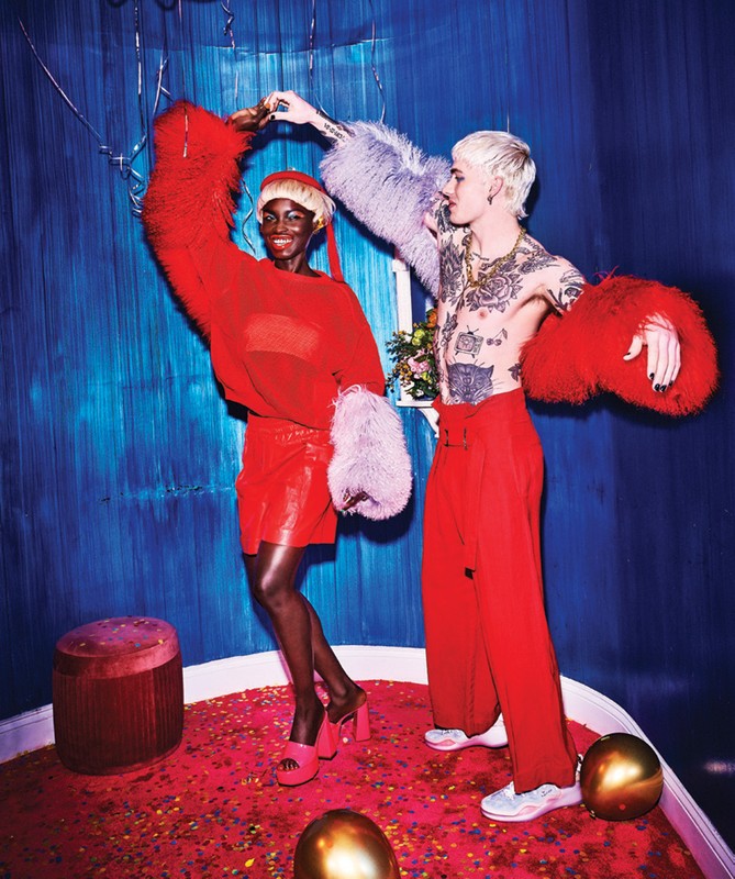 Two fashion models pose in a studio with red carpet and blue backdrop. A black woman in red stands next to a shirtless white man.