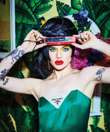 Woman holding a small pink camera to her forehead, wearing a green satin dress and bold makeup with green eyeshadow and red lipstick.
