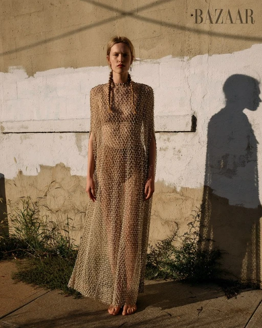 Woman in sheer dress stands outdoors against aged wall, capturing a high-fashion editorial style under natural lighting with 'BAZAAR' visible.