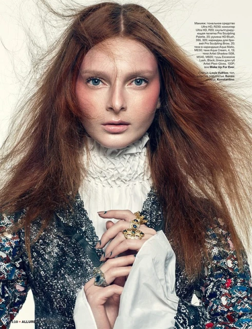 Portrait of a redhead woman with pale skin and pink blush wearing ruff-collared white blouse and ornate jacket with hands near face.