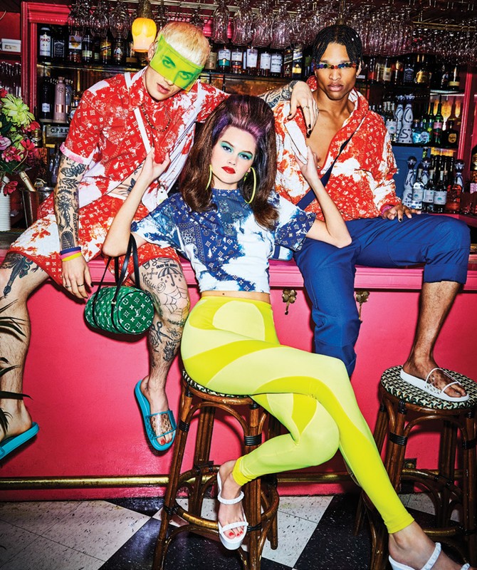 Eye-level shot of three fashion models in patterned shirts and coordinated pants, posing in a retro-styled bar with bright pink walls.
