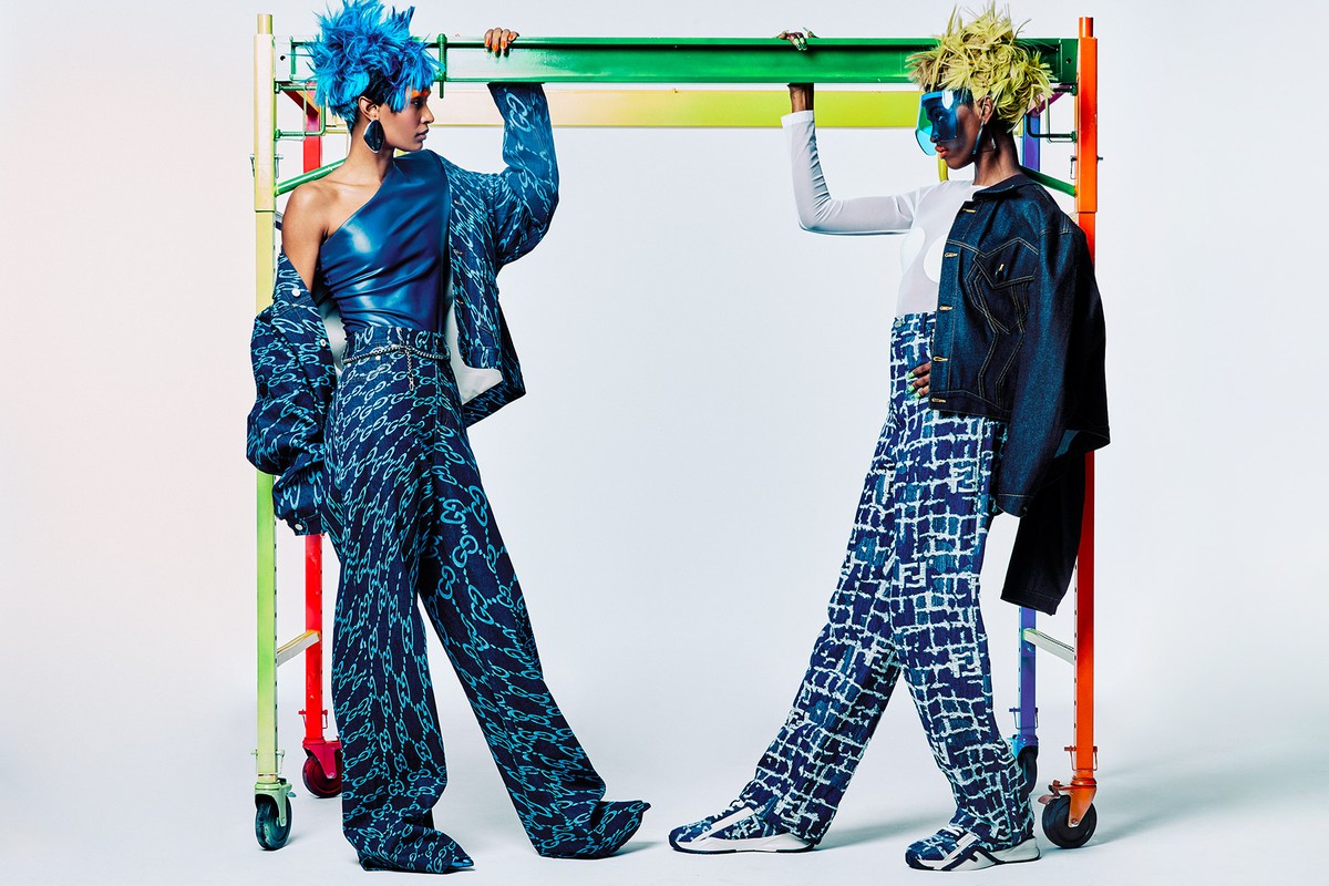 Two models pose in patterned denim outfits, standing in front of a colorful metal scaffolding structure in a studio setting, fashion forward.