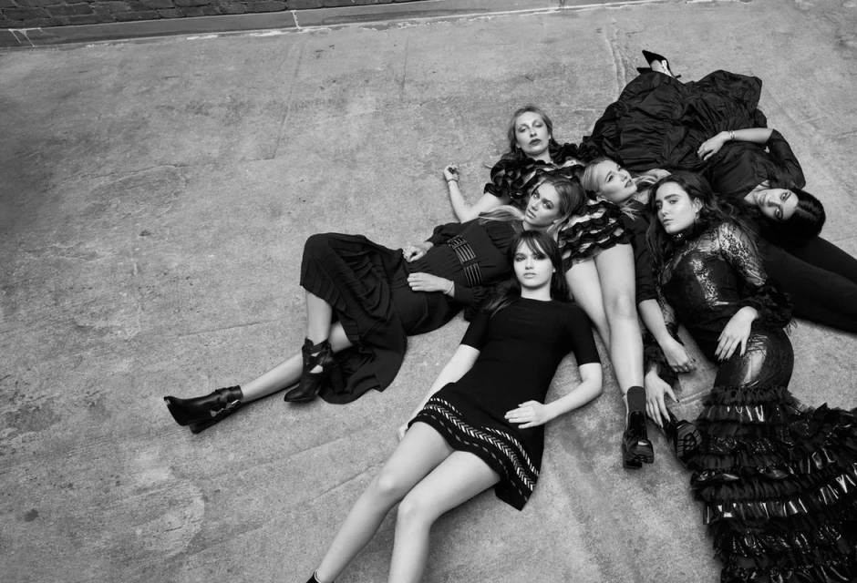 Seven women are lying on a concrete surface wearing various black dresses and black shoes, captured in a black and white, high-angle shot.