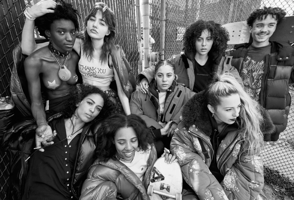Group of seven young adults in an outdoor urban setting, wearing streetwear fashion, some in jackets, and one holding a skateboard.