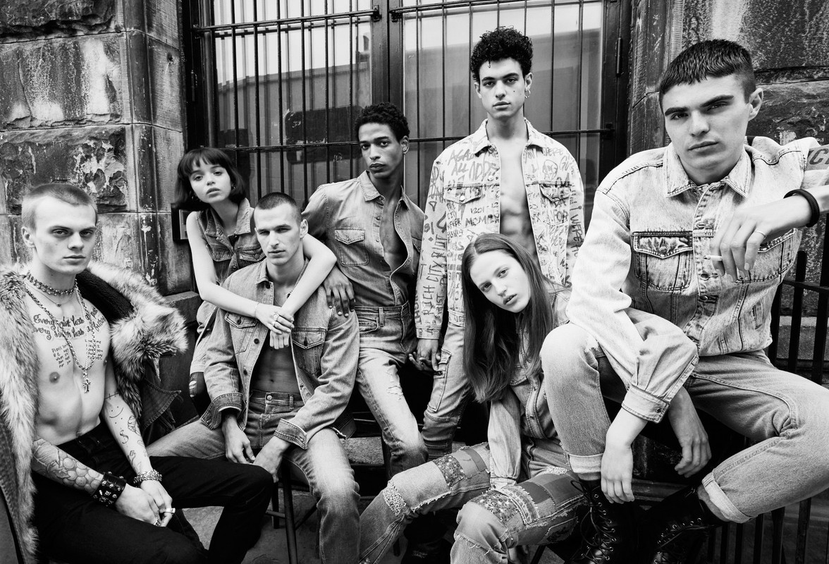 A group of seven young adults models denim clothing in an outdoor urban environment, presented in black and white with a shallow depth of field.