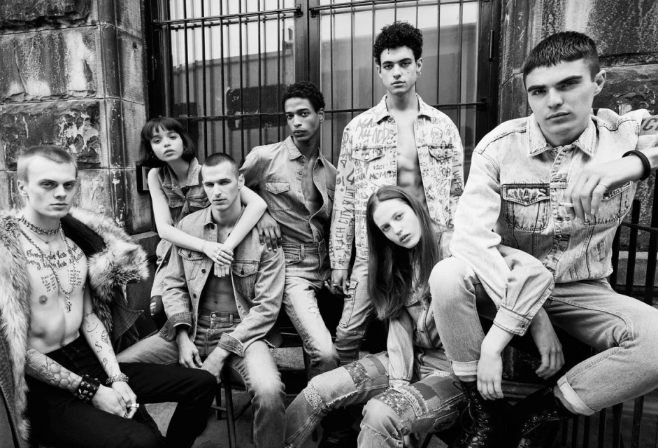 A group of seven young adults models denim clothing in an outdoor urban environment, presented in black and white with a shallow depth of field.
