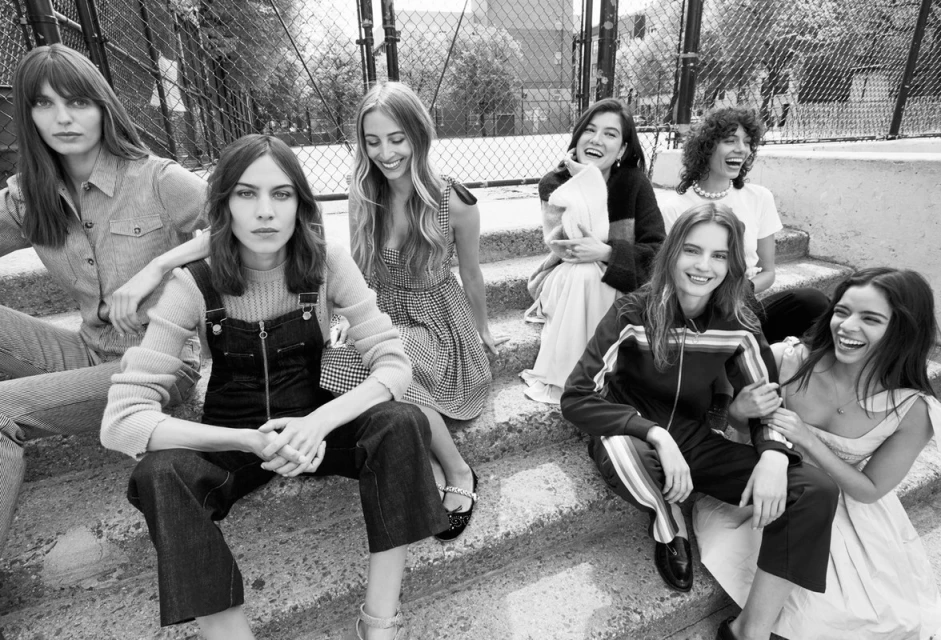 Seven women in varied fashions pose on outdoor steps with a chain-link fence and trees visible in the background in a monochrome shot.