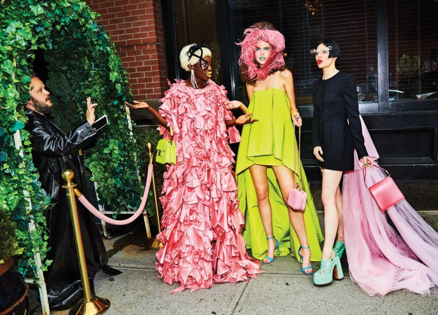 Four fashionably dressed people pose outside a building with greenery. Three women in colorful gowns and heels, and one man photographing them.