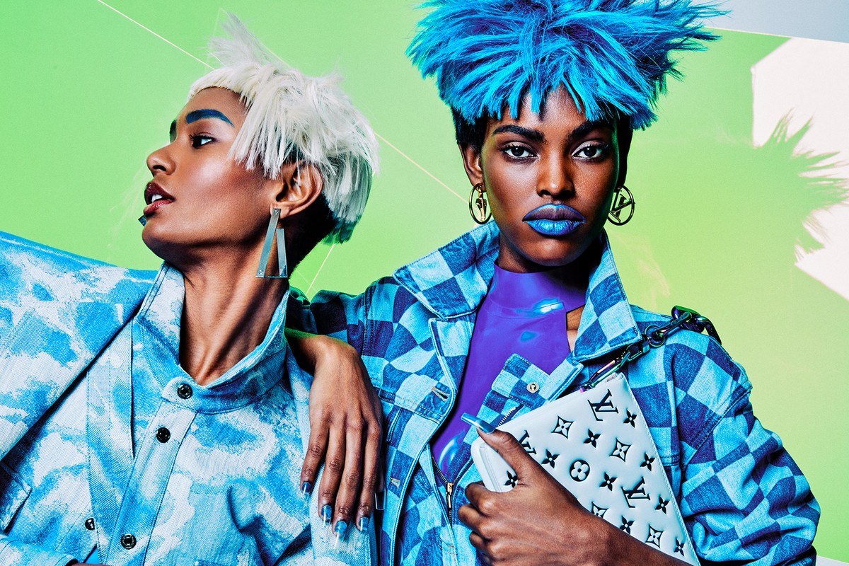 Two fashion models pose in blue-toned outfits with dramatic hair and makeup against a bright green backdrop, accessorized with statement earrings.