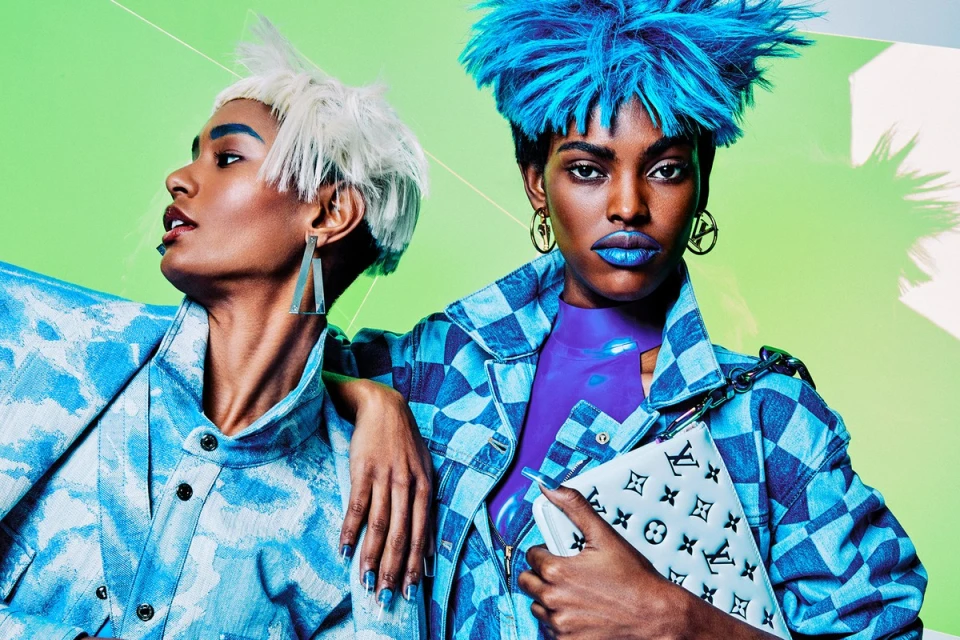 Two fashion models pose in blue-toned outfits with dramatic hair and makeup against a bright green backdrop, accessorized with statement earrings.