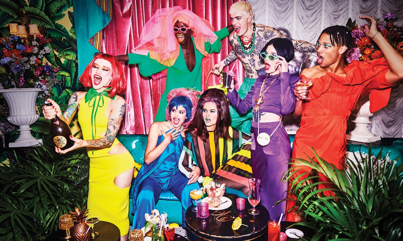 A group of seven diverse people in brightly colored outfits gather around a table in a vibrant, indoor setting with tropical plants.