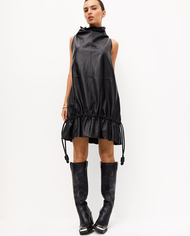 Model in a black leather mini dress and black leather boots standing against a white seamless background