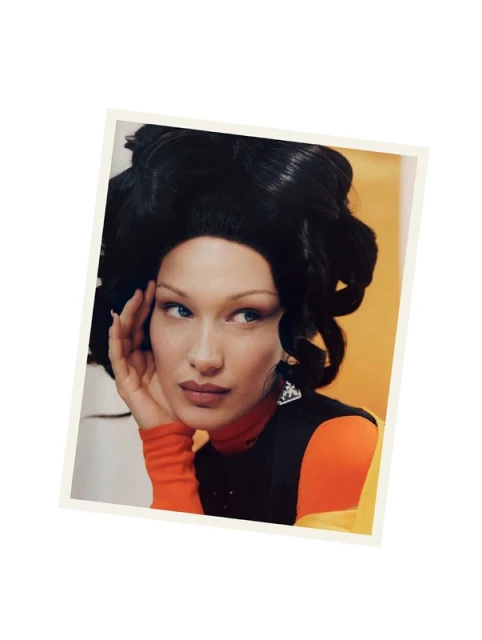 Portrait of model Bella Hadid with elaborate black hair, orange turtleneck and graphic earrings in a fashion studio setting.