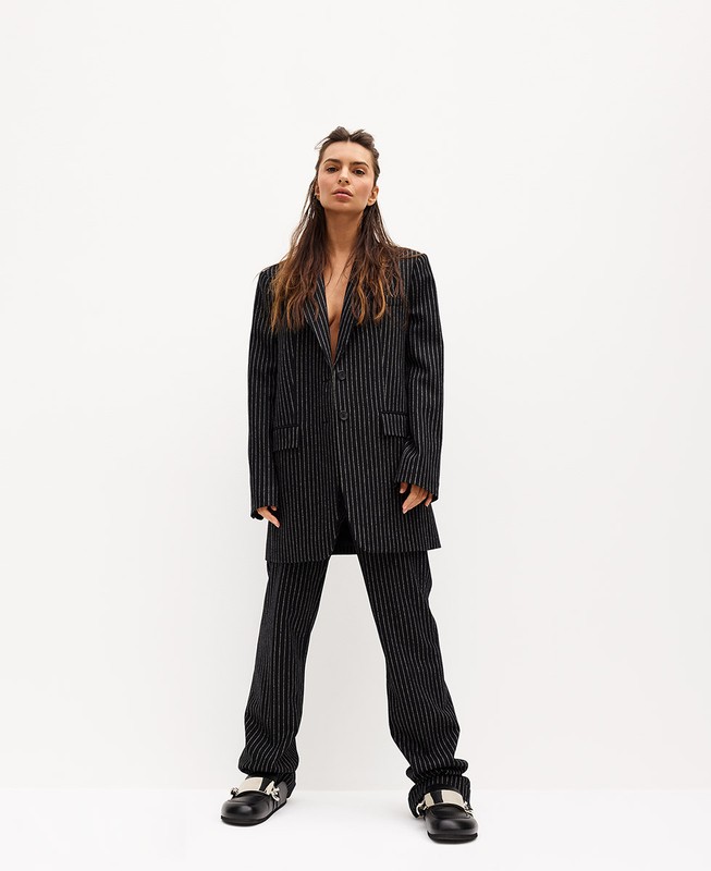 A young adult woman in a pinstripe suit stands against a bright white wall, looking directly at the camera with a neutral expression.