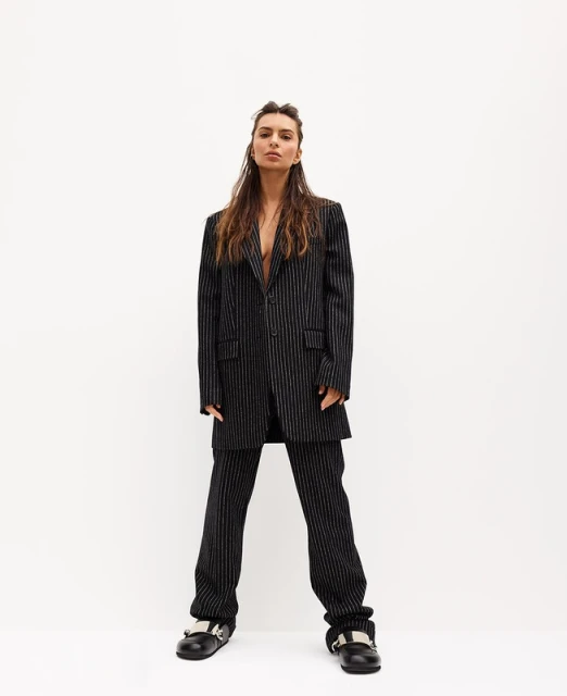 A young adult woman in a pinstripe suit stands against a bright white wall, looking directly at the camera with a neutral expression.